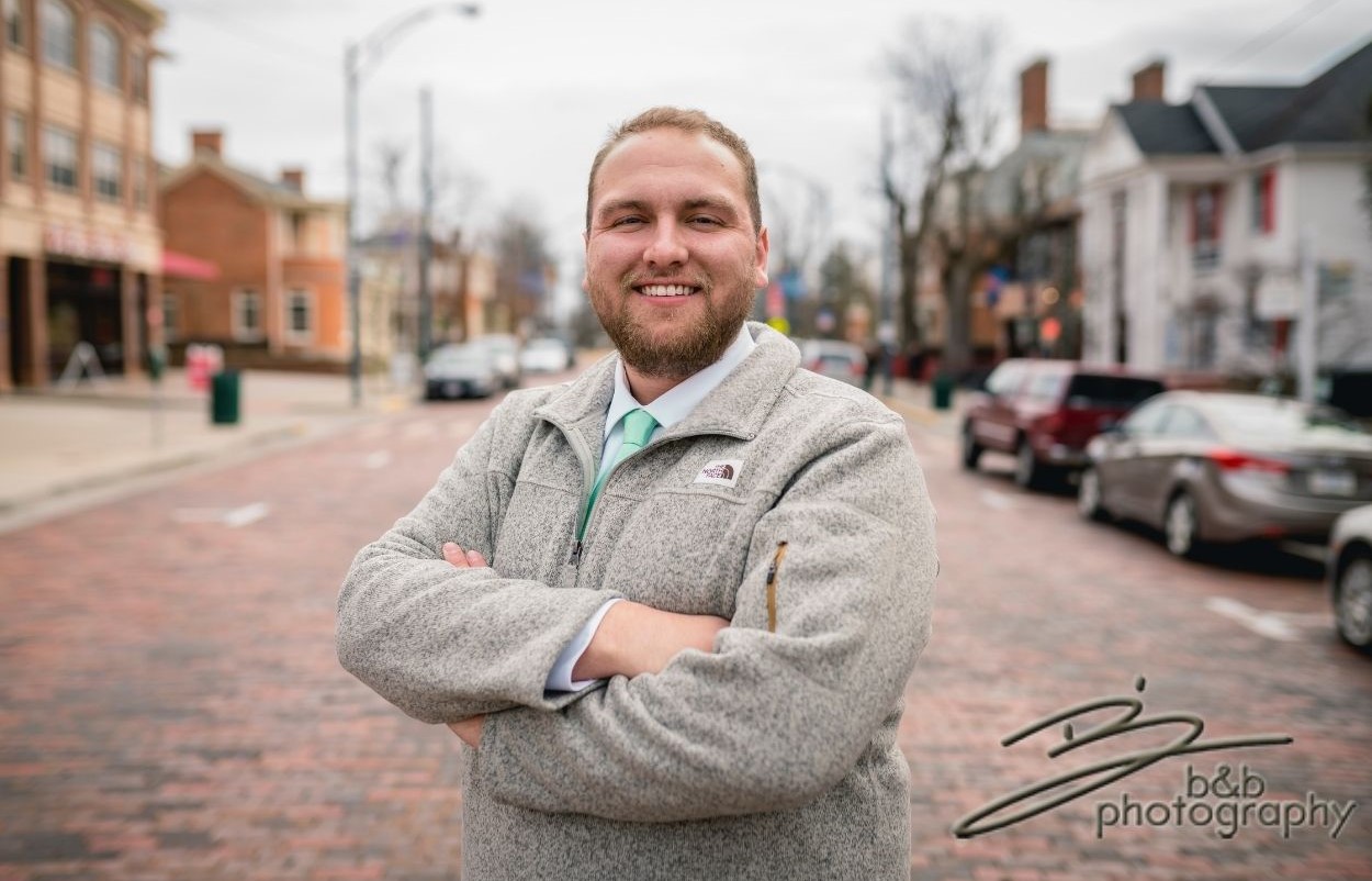 Man posing for a professional headshot in a collared shirt on a brick-covered road.