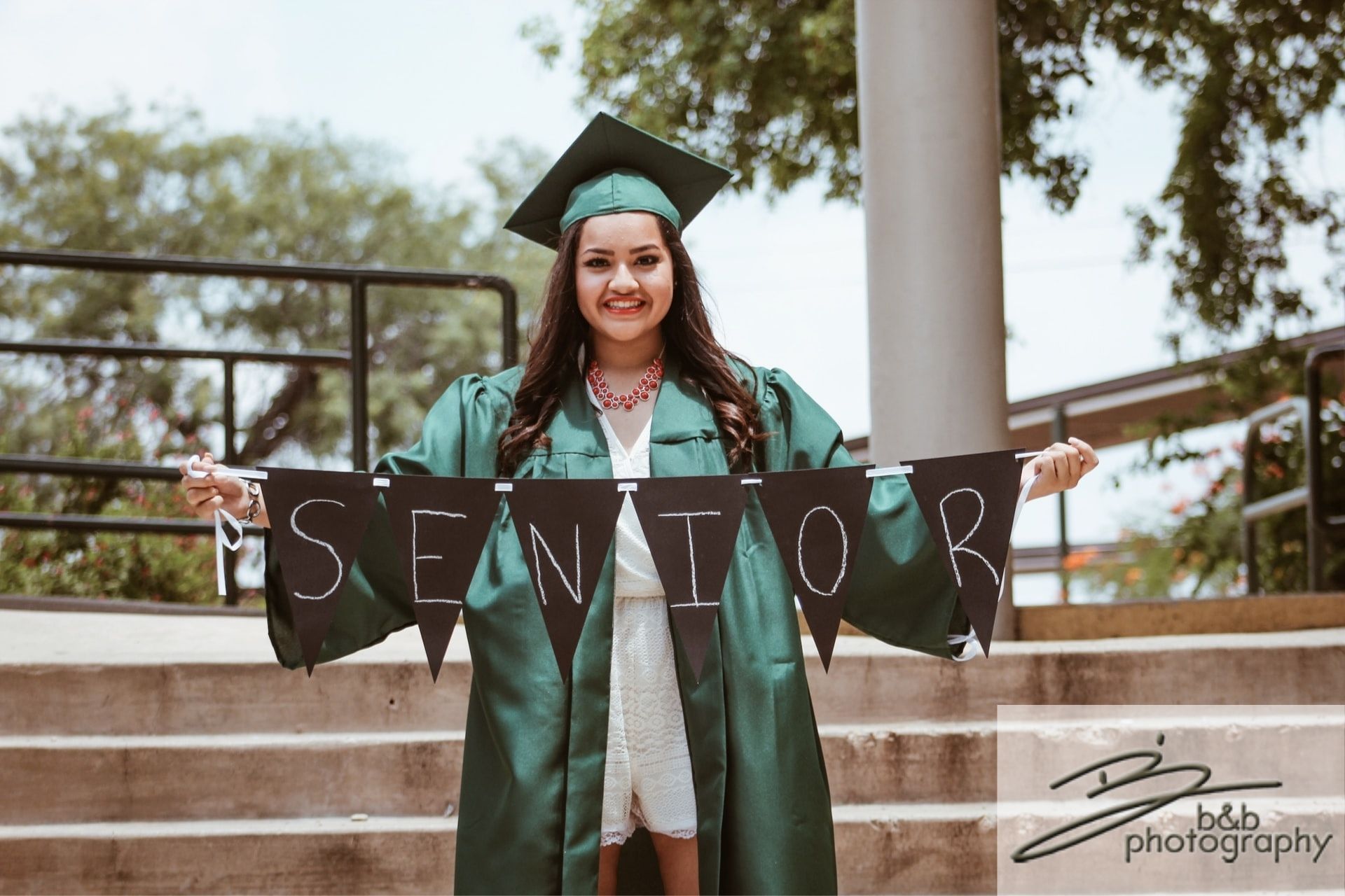 A girl in a white dress with a green cap and gown standing outside holding a senior banner